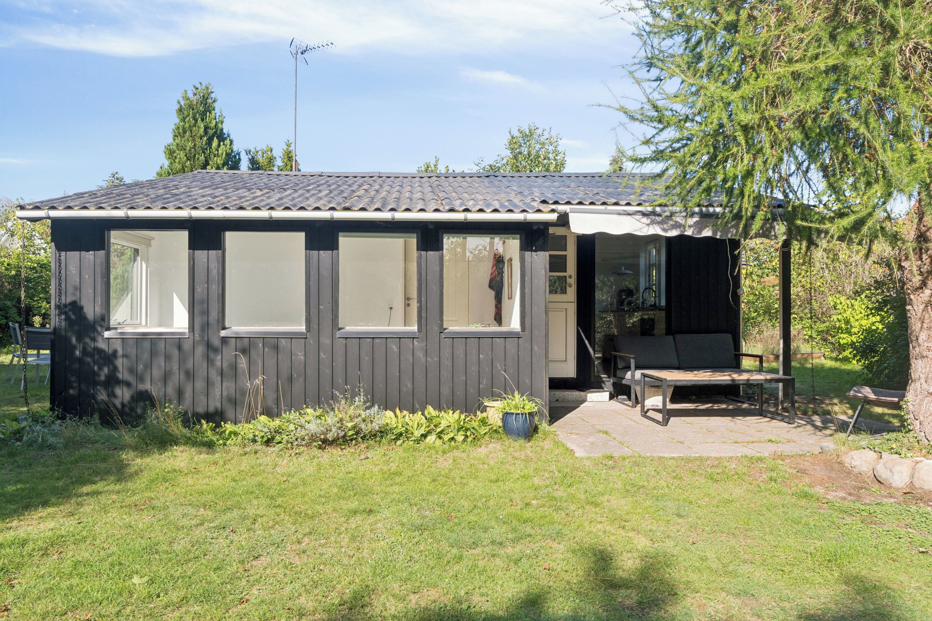 A small, single-story building with a tiled roof, a front porch with a bench, and a small garden area.