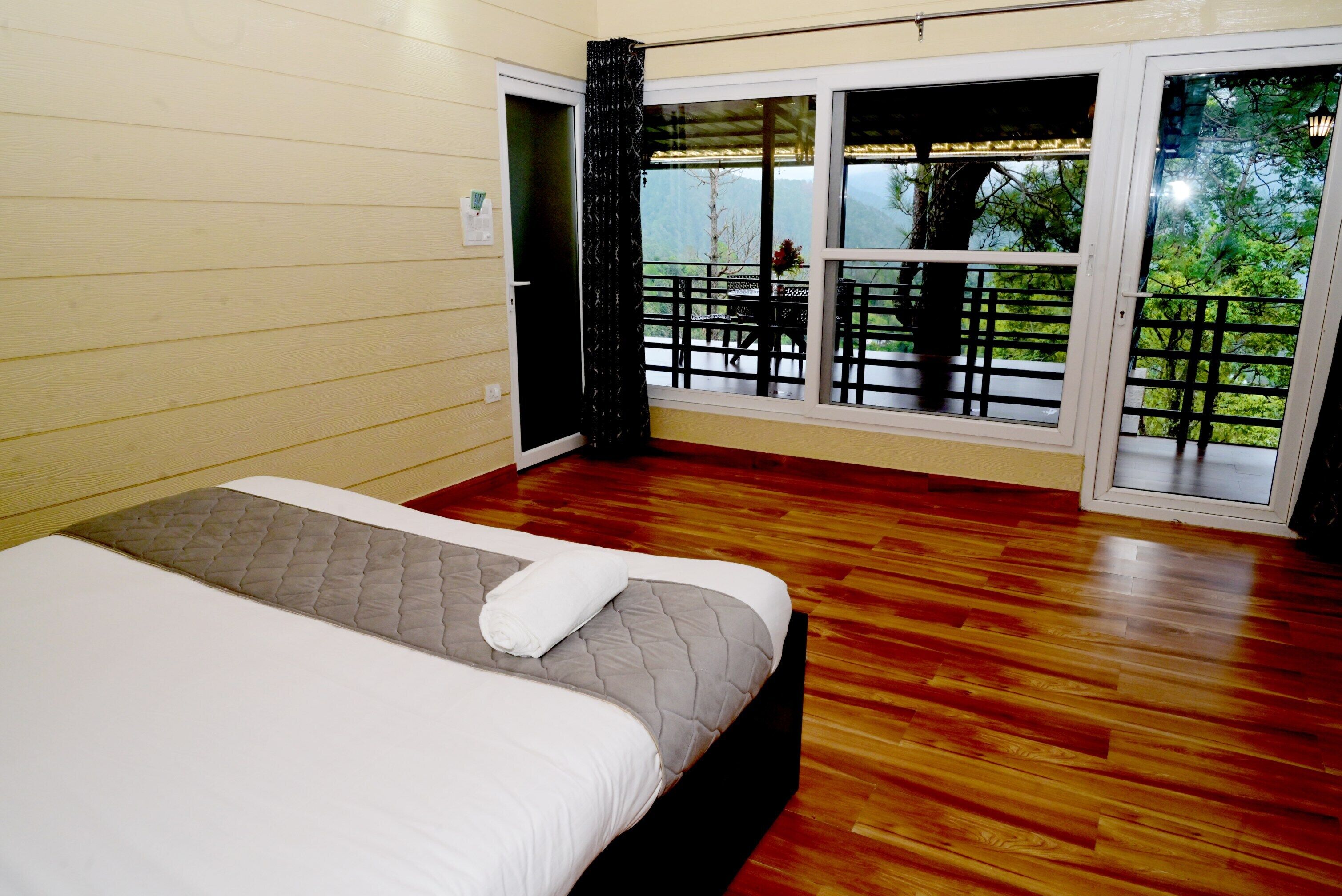 Een slaapkamer met een bed, houten vloeren, een raam met uitzicht op groen en een balkon met buitenmeubilair.