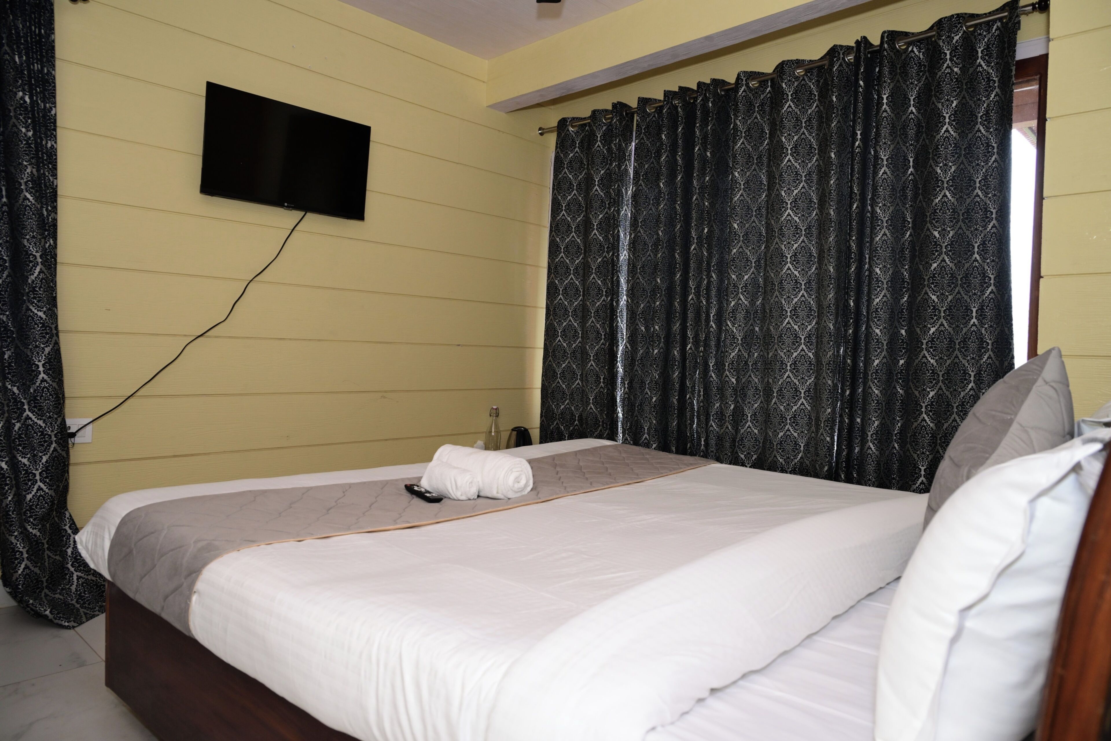 A hotel room with a bed, a TV mounted on the wall, and a window with patterned curtains.