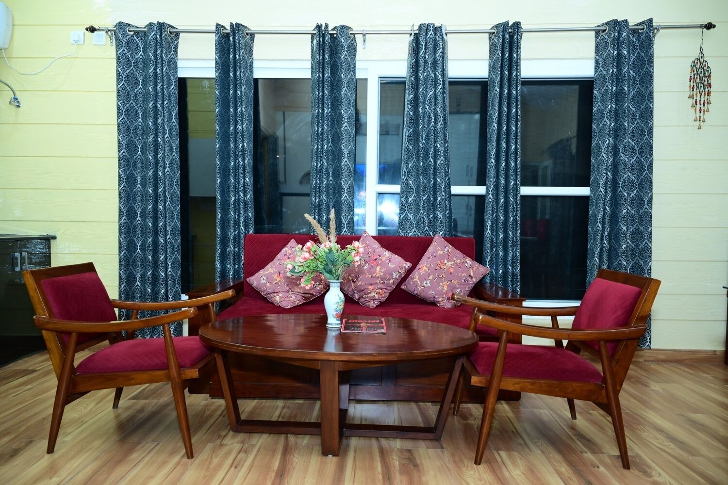 A room with a round wooden table, four wooden chairs with red cushions, a sofa with floral cushions, and patterned curtains.