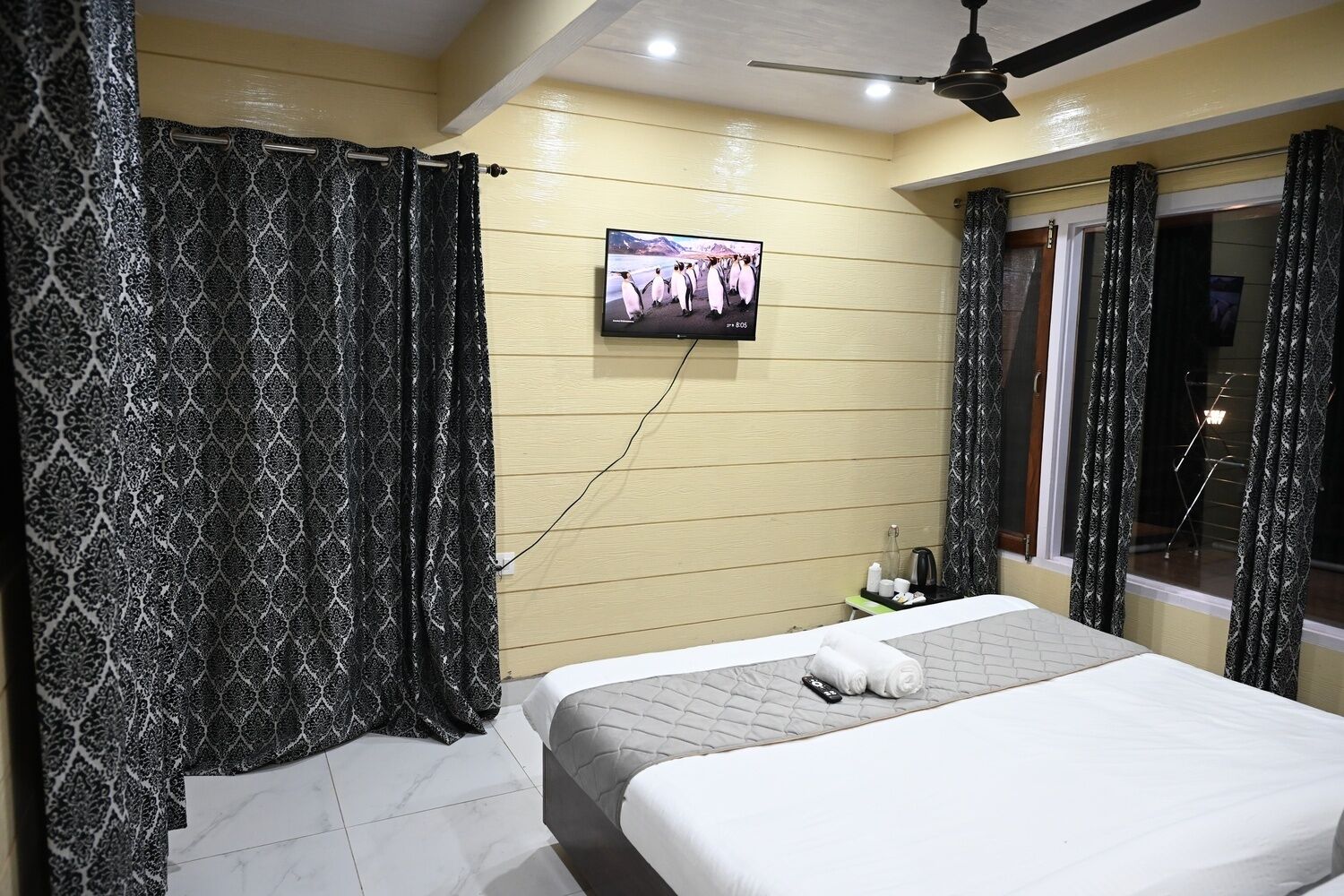 Habitación de hotel con cama, cortinas, televisor montado en la pared y ventana con cortinas.