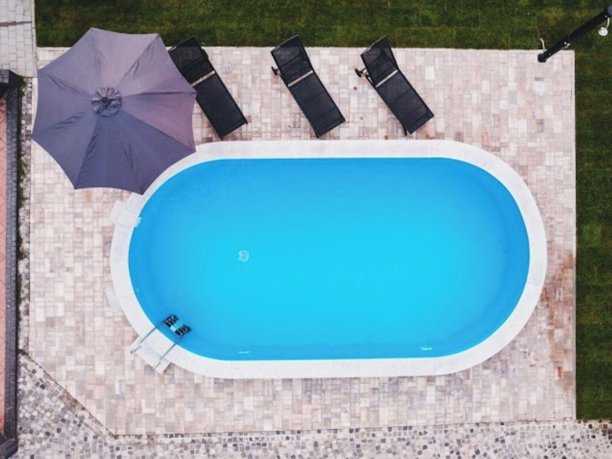 Une piscine rectangulaire avec une terrasse carrelée, une échelle et un espace de détente comprenant des chaises et un parasol.