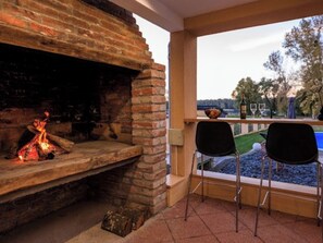 A brick fireplace with a fire, a pool, and two chairs outside.