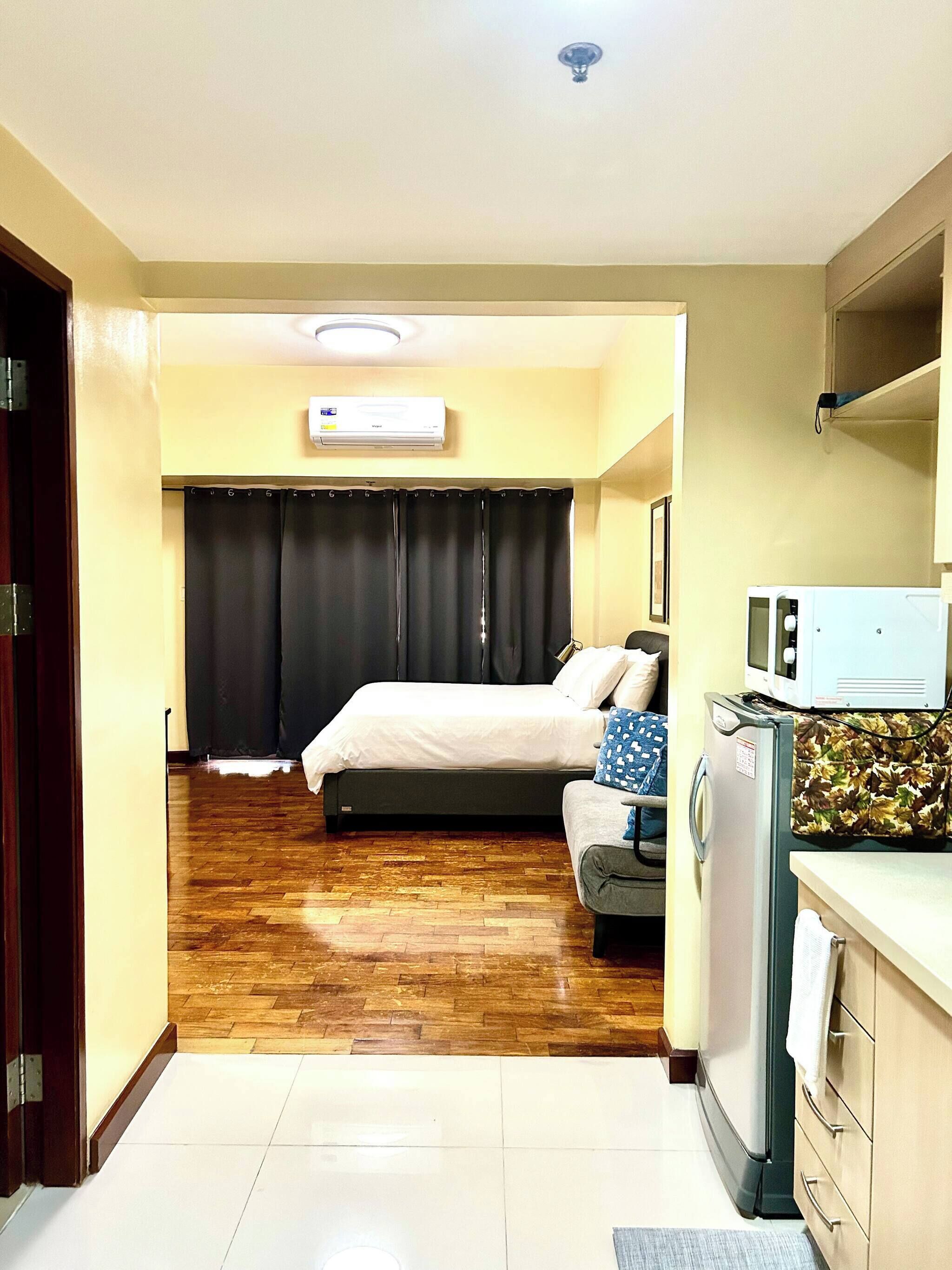 A hotel room with a bed, a sofa, a microwave, a refrigerator, and an air conditioning unit.
