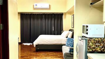 A hotel room with a bed, a sofa, a microwave, a refrigerator, and an air conditioning unit.