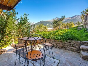 Une terrasse extérieure avec une table en pierre et des chaises en métal, entourée d’une végétation luxuriante et offrant une vue sur les montagnes.