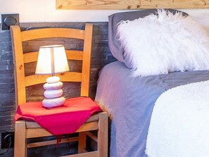 En træstol med en lampe og en rød pude, en seng med en hvid pude, og en stenvæg.