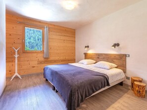 A bedroom with a wooden headboard, a bed with white bedding, a window with a view of trees, and a wooden floor. - Chalet Cairn by Interhome (Peisey-Nancroix)