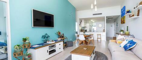 Una sala de estar moderna con una pared de acento color turquesa, un televisor de pantalla plana, un área de comedor y una cocina al fondo, además de un sofá con cojines decorativos.