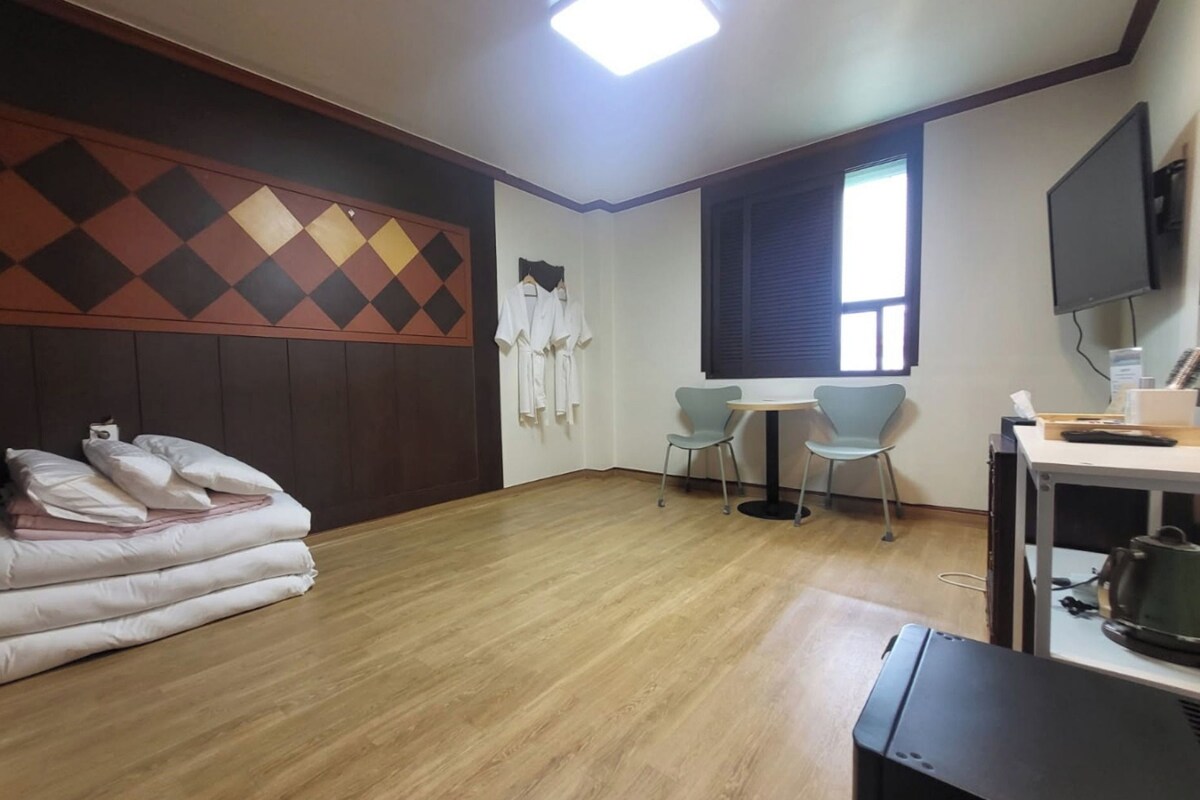 Una habitación con cama, una mesita con sillas, televisor y ventana con persianas.