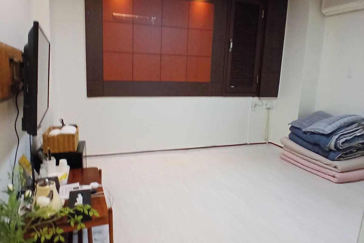 Una habitación con una mesa de madera, un televisor en la pared, una ventana con persianas y una pila de ropa doblada.