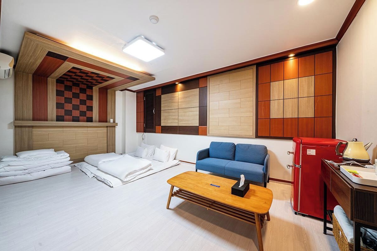 Una habitación con una cama, un sofá azul, una mesa de centro de madera, un refrigerador rojo y una pared con estampado de cuadros.