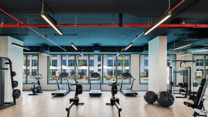 Un gimnasio con cintas de correr, elípticas y equipo de pesas, amplias ventanas y vista a mesas al aire libre.