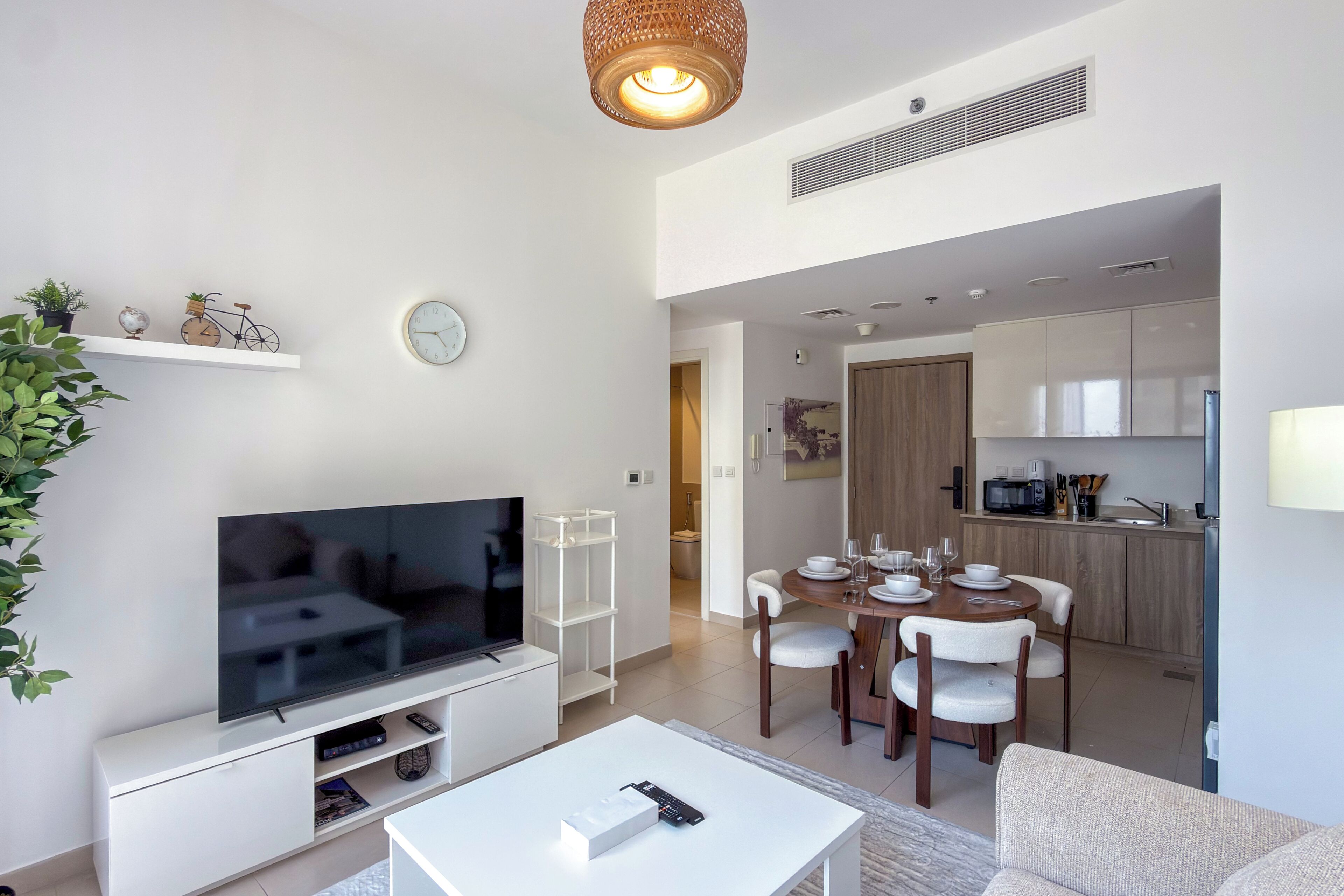 A modern living room with a flat-screen TV, a dining area with a wooden table and chairs, and a kitchen in the background.