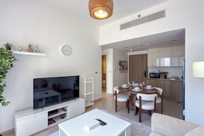 A modern living room with a flat-screen TV, a dining area with a wooden table and chairs, and a kitchen in the background. - UNA Apartments - Town Square Dubai (Dubai)