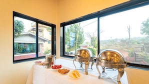 Ein reich gedeckter Büfett mit Speisen und silbernen Serviergeschirr auf einem Tisch, mit Fenstern, die einen Blick auf Gebäude und Grünflächen bieten.