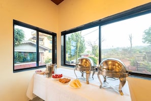 Mesa con una barra de buffet, alimentos y vajilla de plata, y ventanas con vistas a edificios y áreas verdes.