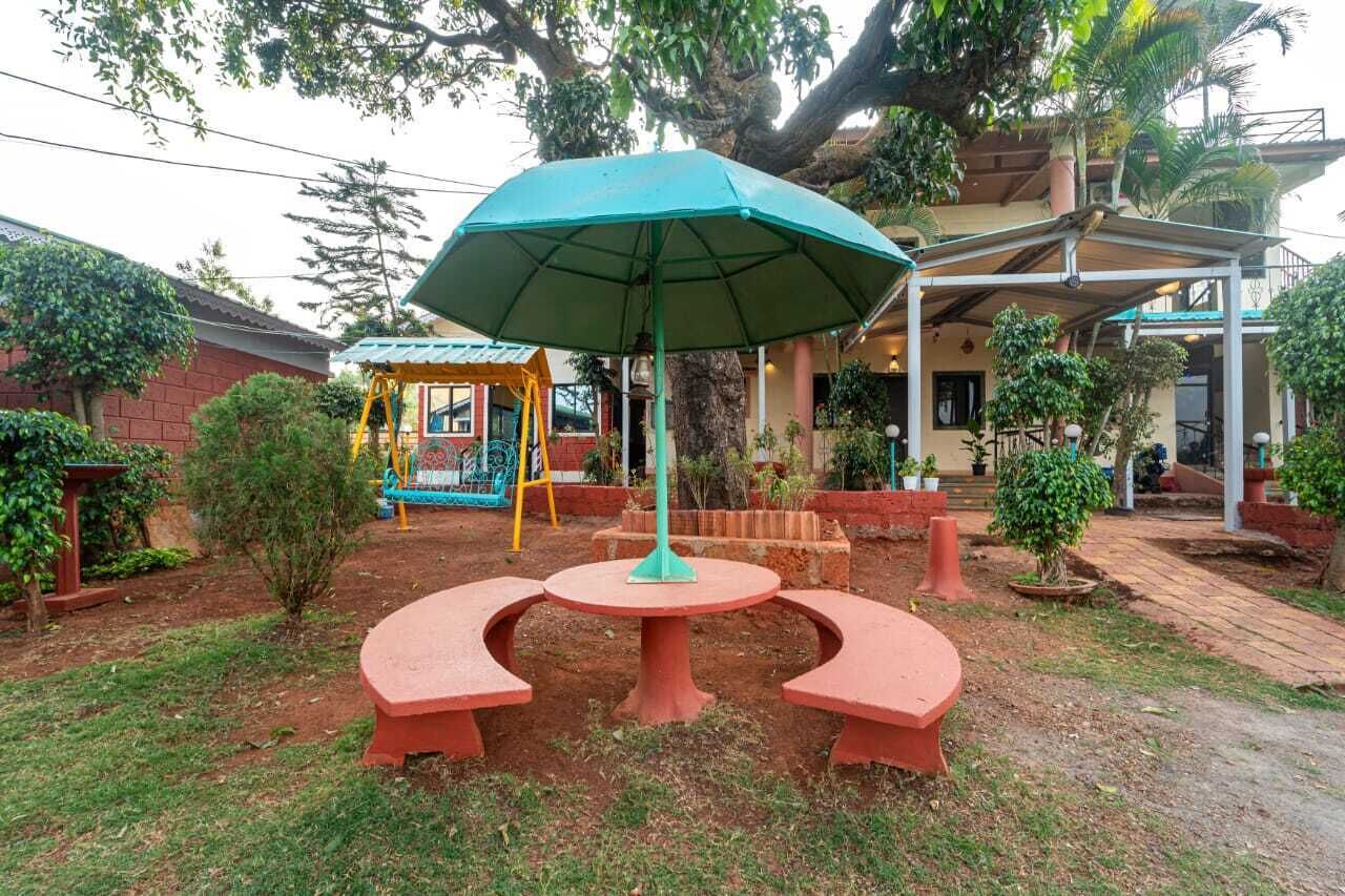 An outdoor seating area with a table and benches, surrounded by greenery and a building in the background.