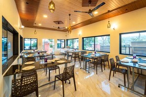 A restaurant with multiple tables and chairs, wooden ceiling, and large windows. - Hiltan Valley Resort (Mahabaleshwar)