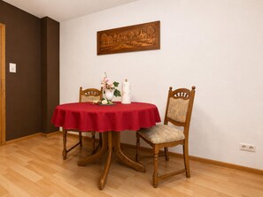 Un comedor con una mesa redonda cubierta por un mantel rojo, dos sillas de madera, un florero con flores y una obra de arte colgada en la pared.