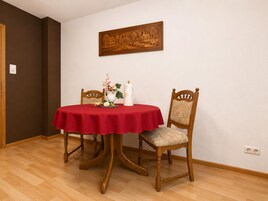 Ein Essbereich mit einem runden Tisch, der mit einer roten Tischdecke bedeckt ist, zwei Holzstühlen, einer Blumenvase und einem an der Wand hängenden Kunstwerk.