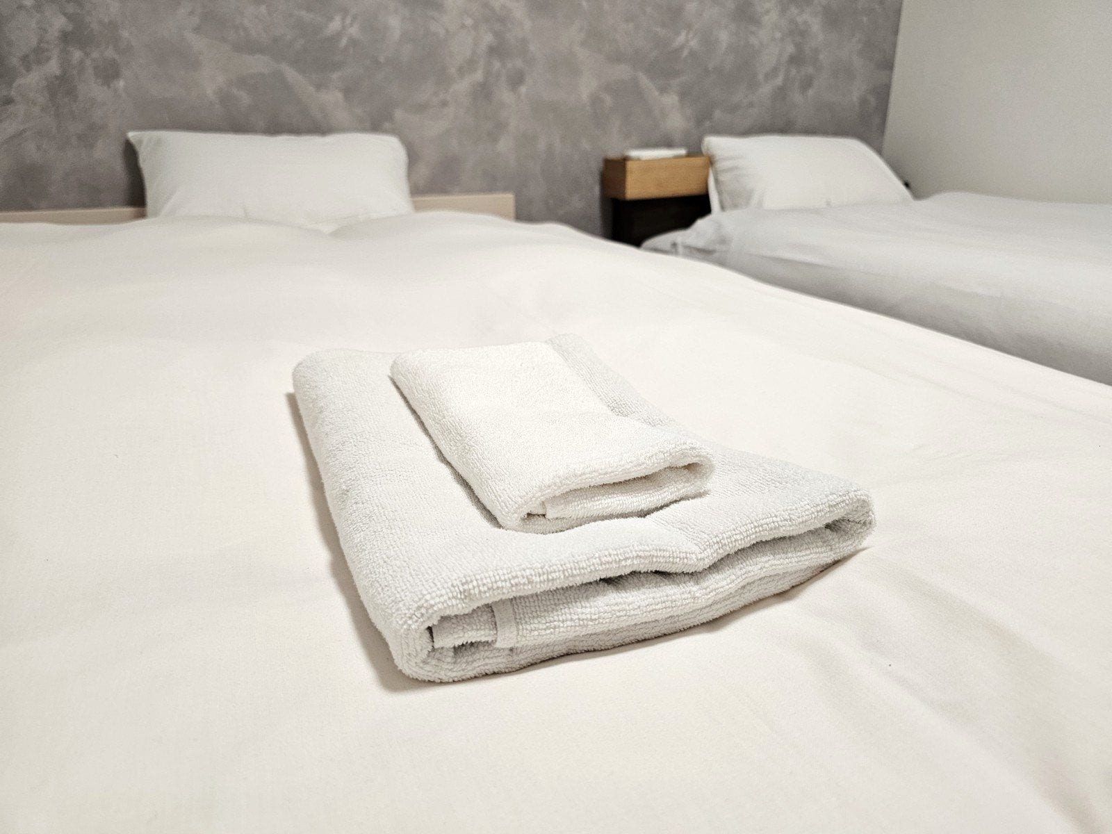A hotel room with two beds, each with white linens and pillows, and a neatly folded white towel on the bed.