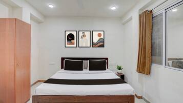 Quarto de hotel com uma cama, um guarda-roupa, uma janela com cortinas e duas molduras na parede.