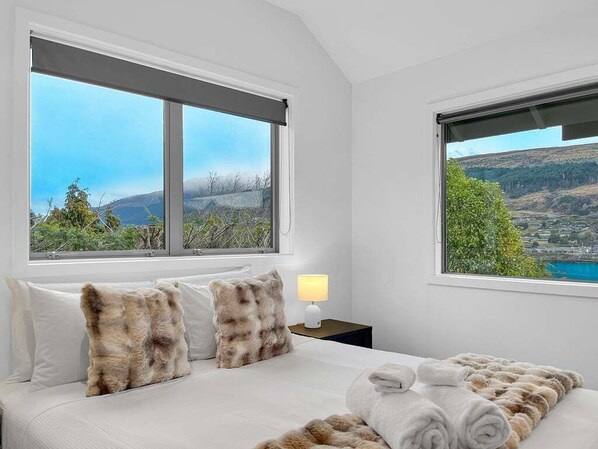 A hotel room with a bed, two windows with views, a nightstand with a lamp, and a rolled towel on the bed.