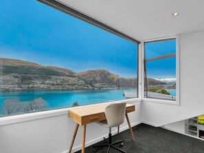 A room with a desk and chair, large windows with a view of a lake and mountains.