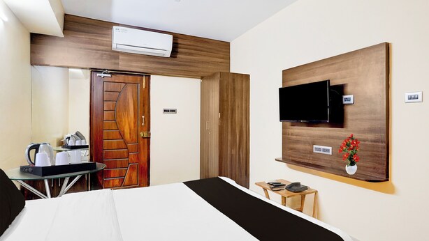 A hotel room with a bed, a TV mounted on the wall, a coffee maker, and a small table with a phone and remote controls.