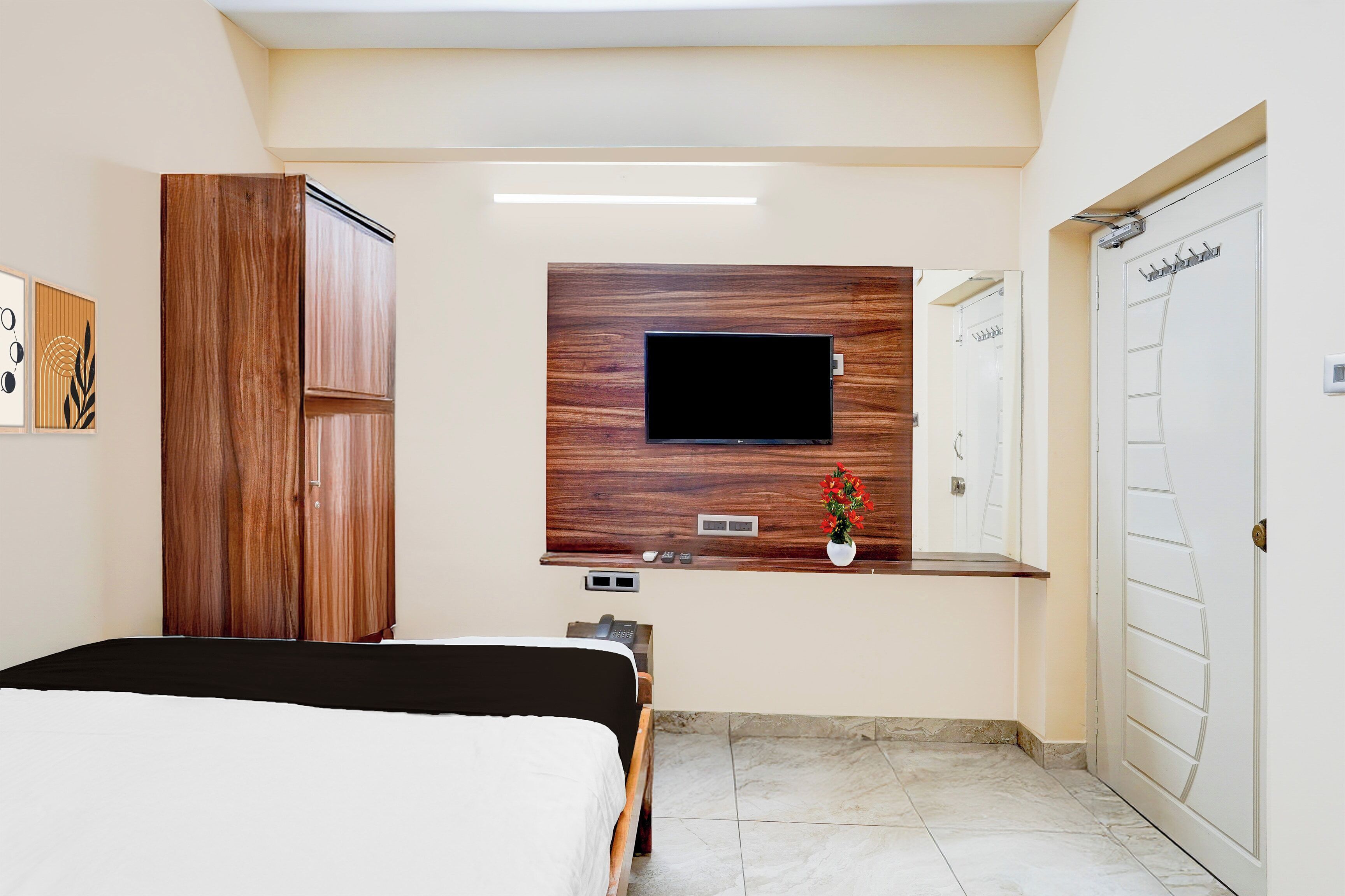 A hotel room with a bed, a wooden wardrobe, a flat-screen TV mounted on a wooden panel, a small desk with a telephone, and a bathroom with a white door.