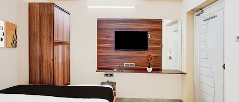 A hotel room with a bed, a wooden wardrobe, a flat-screen TV mounted on a wooden panel, a small desk with a telephone, and a bathroom with a white door.