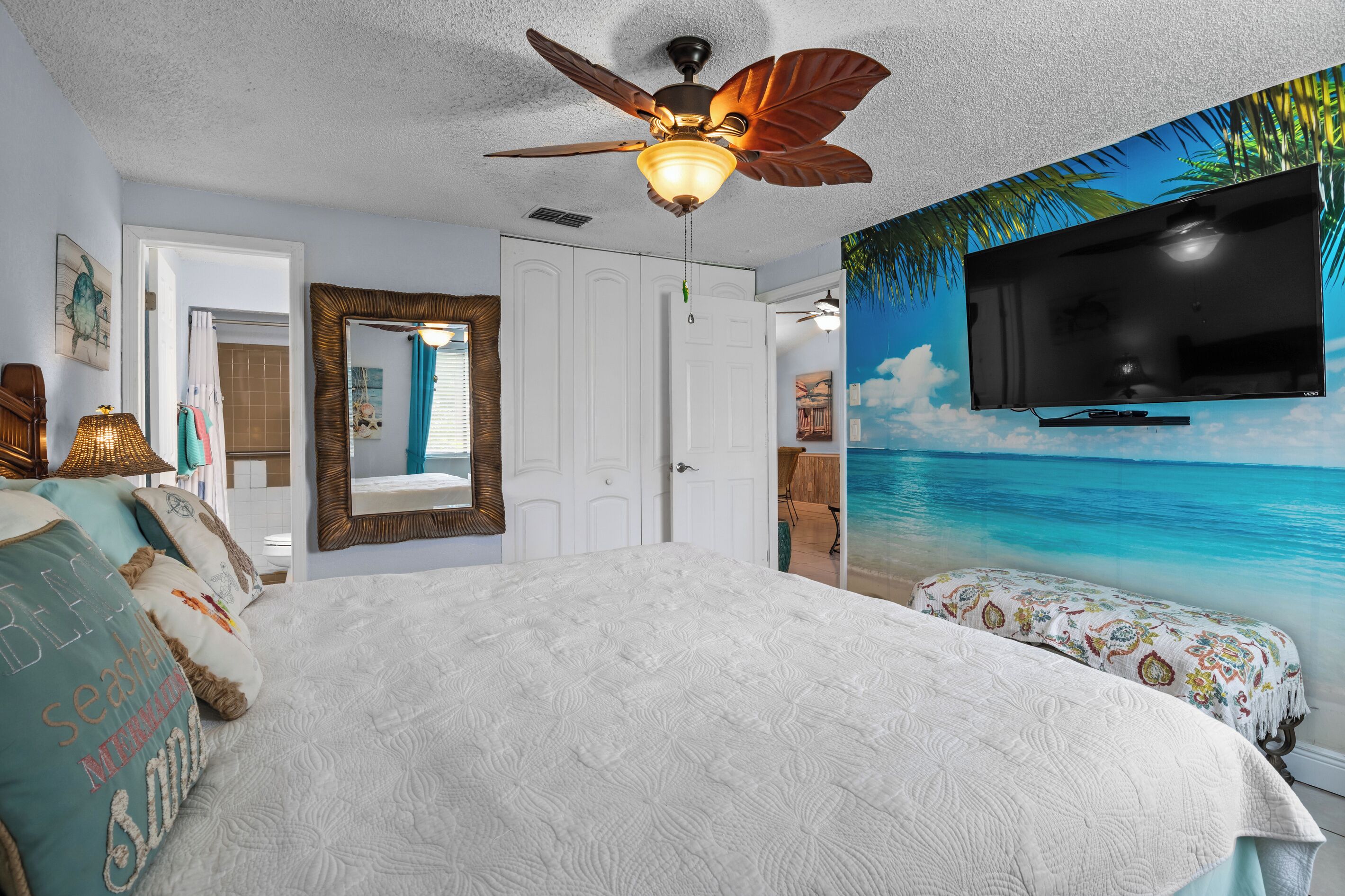 A bedroom with a bed, a ceiling fan, a TV, and a mural of a beach scene.