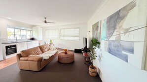 En moderne stue med en sofa, et sofabord og et fjernsyn. Der er et køkkenområde med skabe og et vindue.