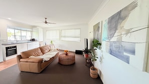Ein modernes Wohnzimmer mit einer Couch, einem Couchtisch und einem Fernseher. Es gibt eine Küchenzeile mit Schränken und einem Fenster.