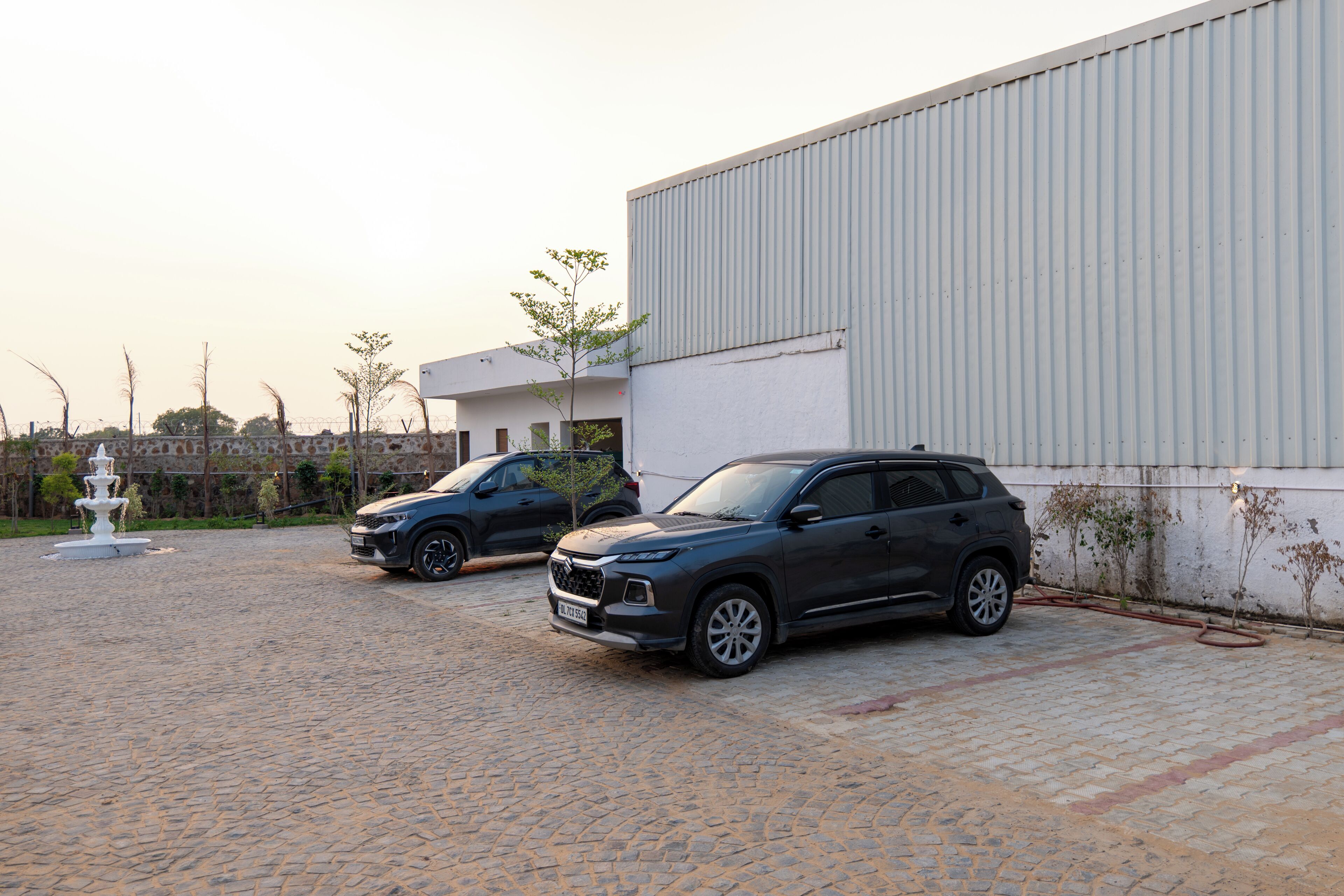 Ein gepflasterter Weg mit zwei geparkten Geländewagen, ein weißes Gebäude mit Wellblechfassade und eine Fontäne im Vordergrund.