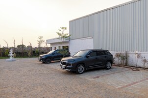 Un camino de entrada pavimentado con dos camionetas estacionadas, un edificio blanco con fachada de metal corrugado y una fuente al frente.