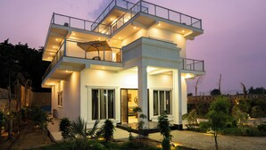 A modern two-story house with a balcony, large windows, and a well-lit entrance. - ELIVAAS JP Farm Stay 5BHK Villa With Bar (Baghanki)