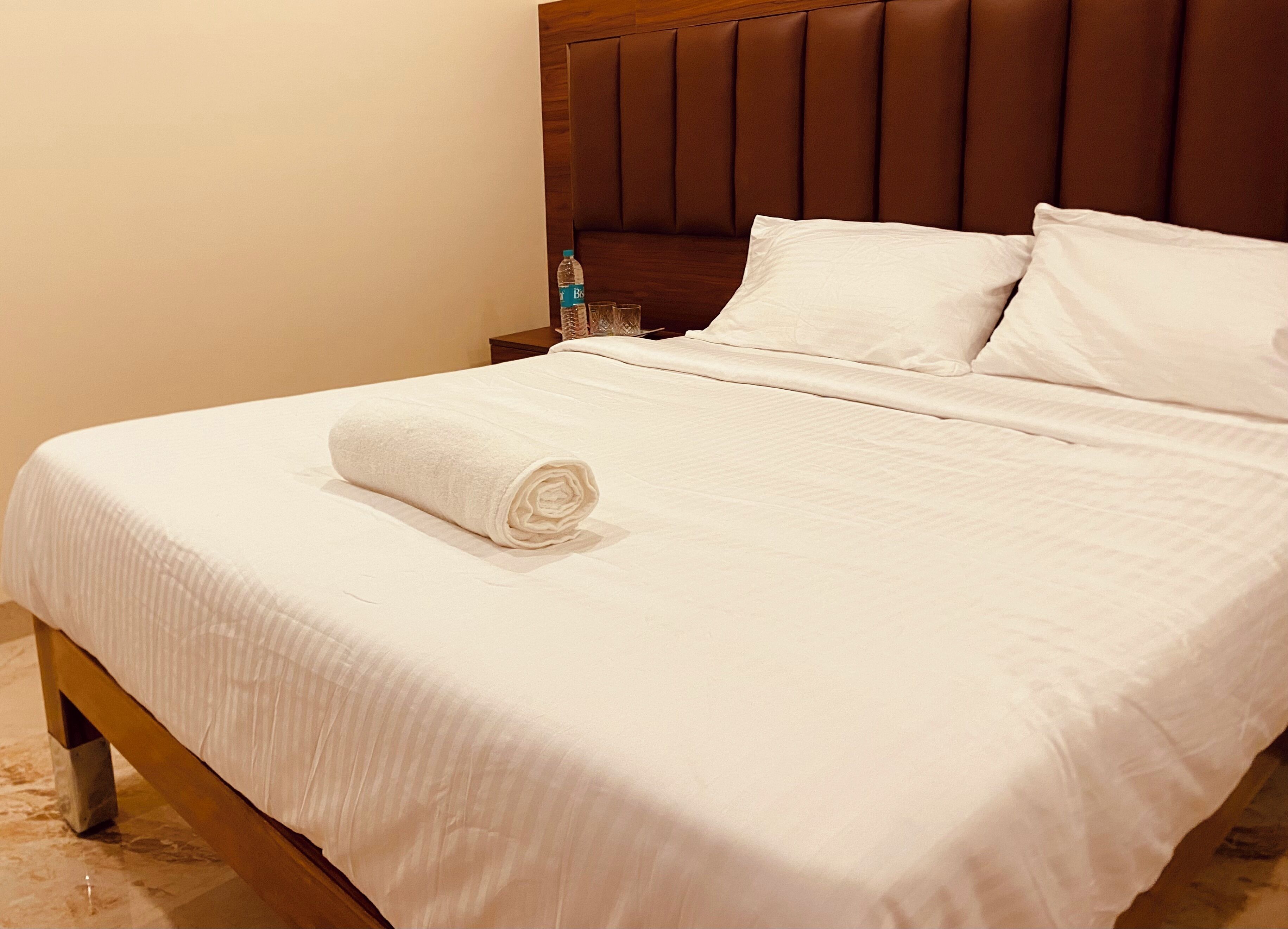 A neatly made bed with a rolled white towel, a wooden headboard, and a nightstand with a bottle and glasses.