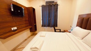 A hotel room with a bed, a wooden headboard, a mounted TV, a window with curtains, and a small table with a bottle.