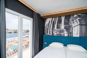 Ein Hotelzimmer mit einem großen Fenster, einem Bett mit weißer Bettwäsche und einem Wandbild eines historischen Gebäudes.