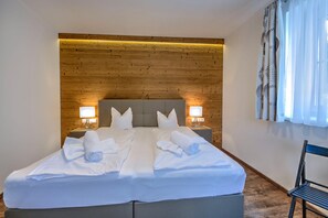 Una cama bien hecha con ropa blanca y toallas, cabecera de madera, dos lámparas de noche y una silla a la derecha.