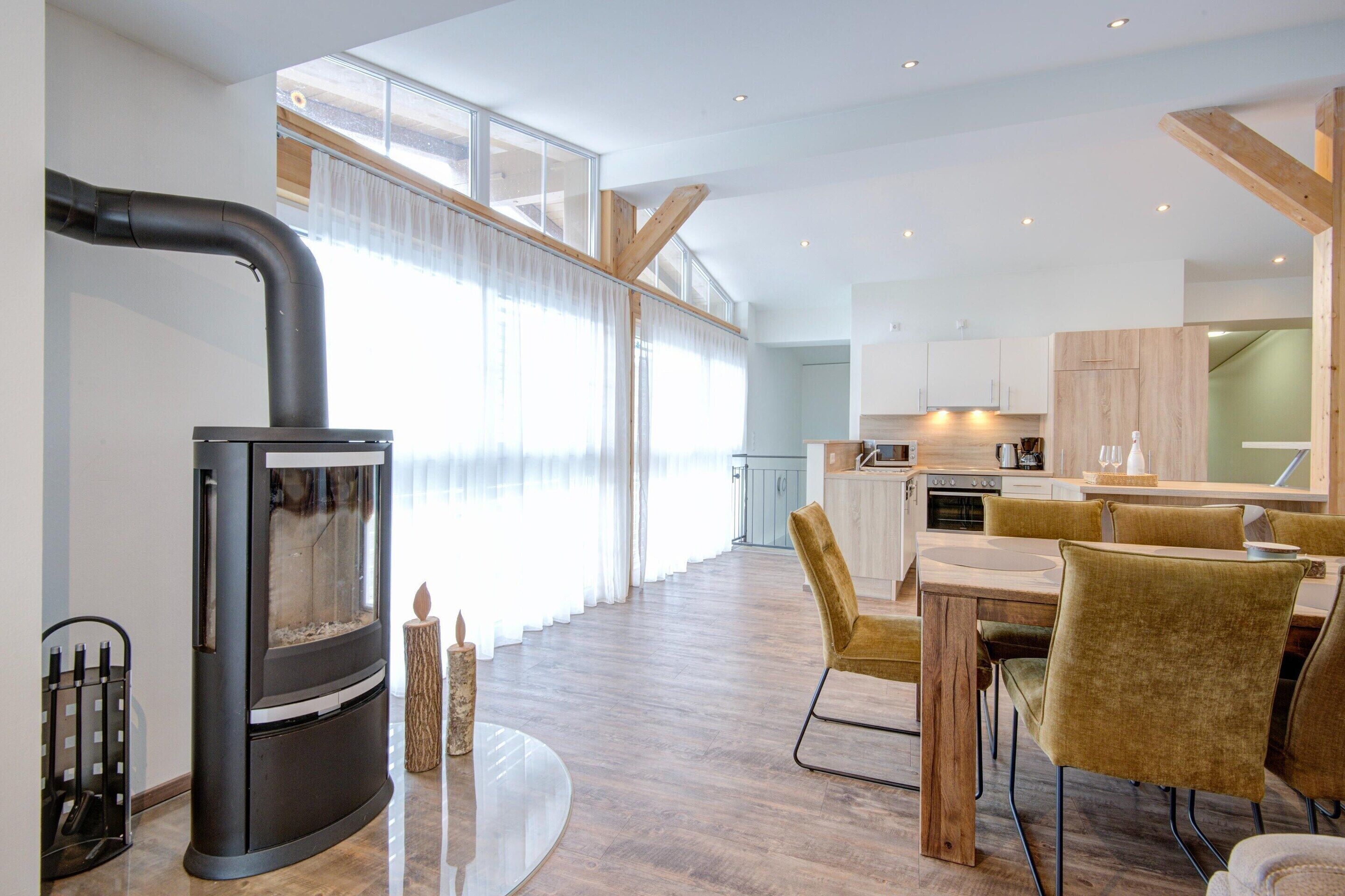 A modern kitchen with wooden cabinets, a dining area with a round table and chairs, and a large window with sheer curtains.