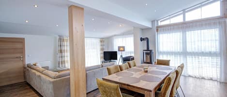 A modern living room with a wooden dining table, comfortable seating, a television, and large windows.