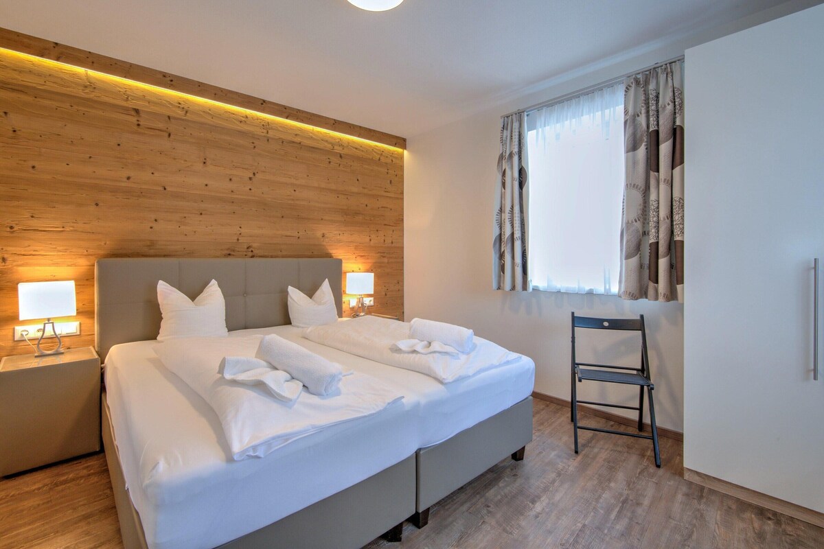 A double bed with white linens and pillows, a wooden headboard, two bedside tables with lamps, a window with curtains, and a chair.
