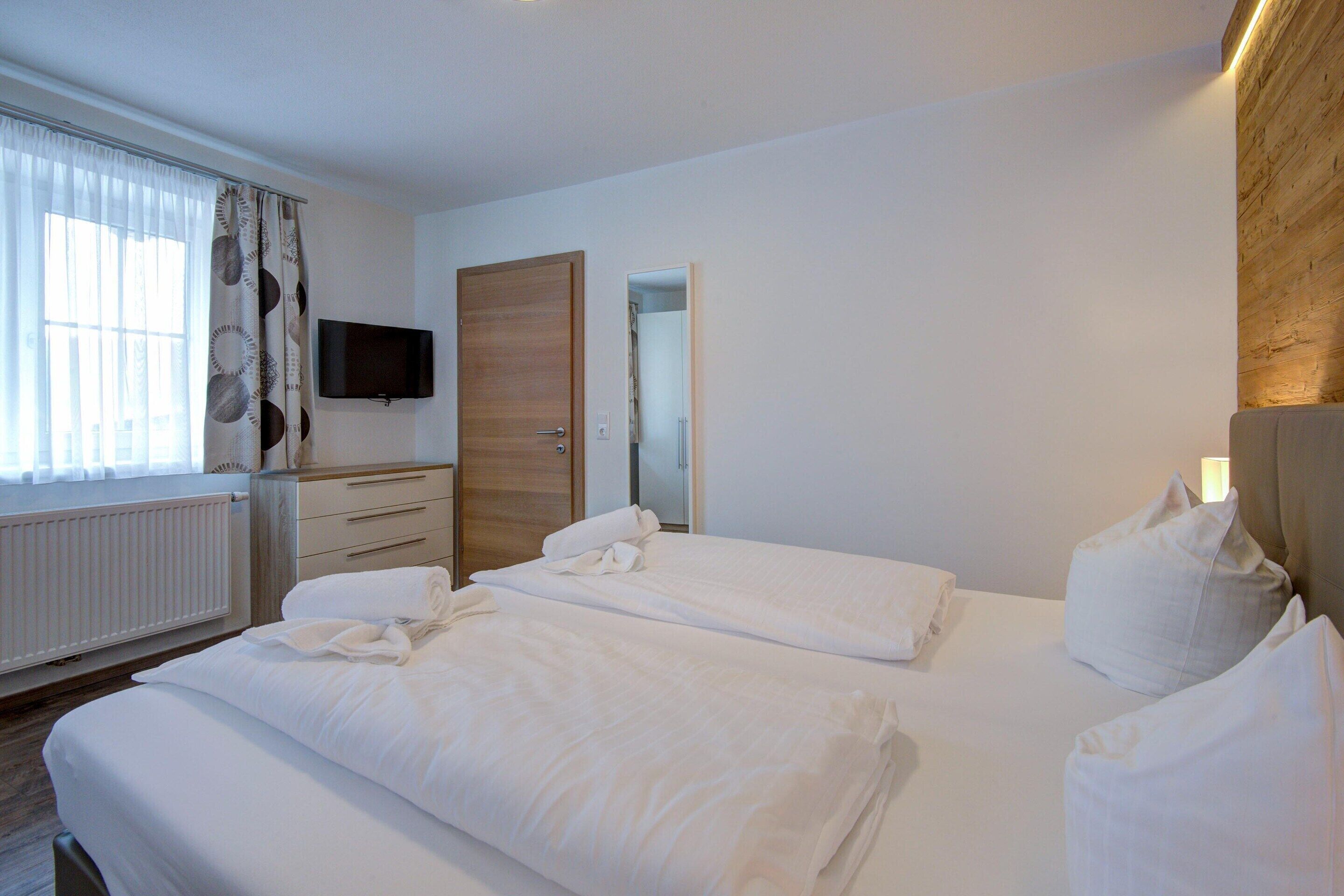Ein Hotelzimmer mit zwei Betten, einem Fernseher, einem hölzernen Kopfteil, einem Spiegel und einem Fenster mit Vorhängen.