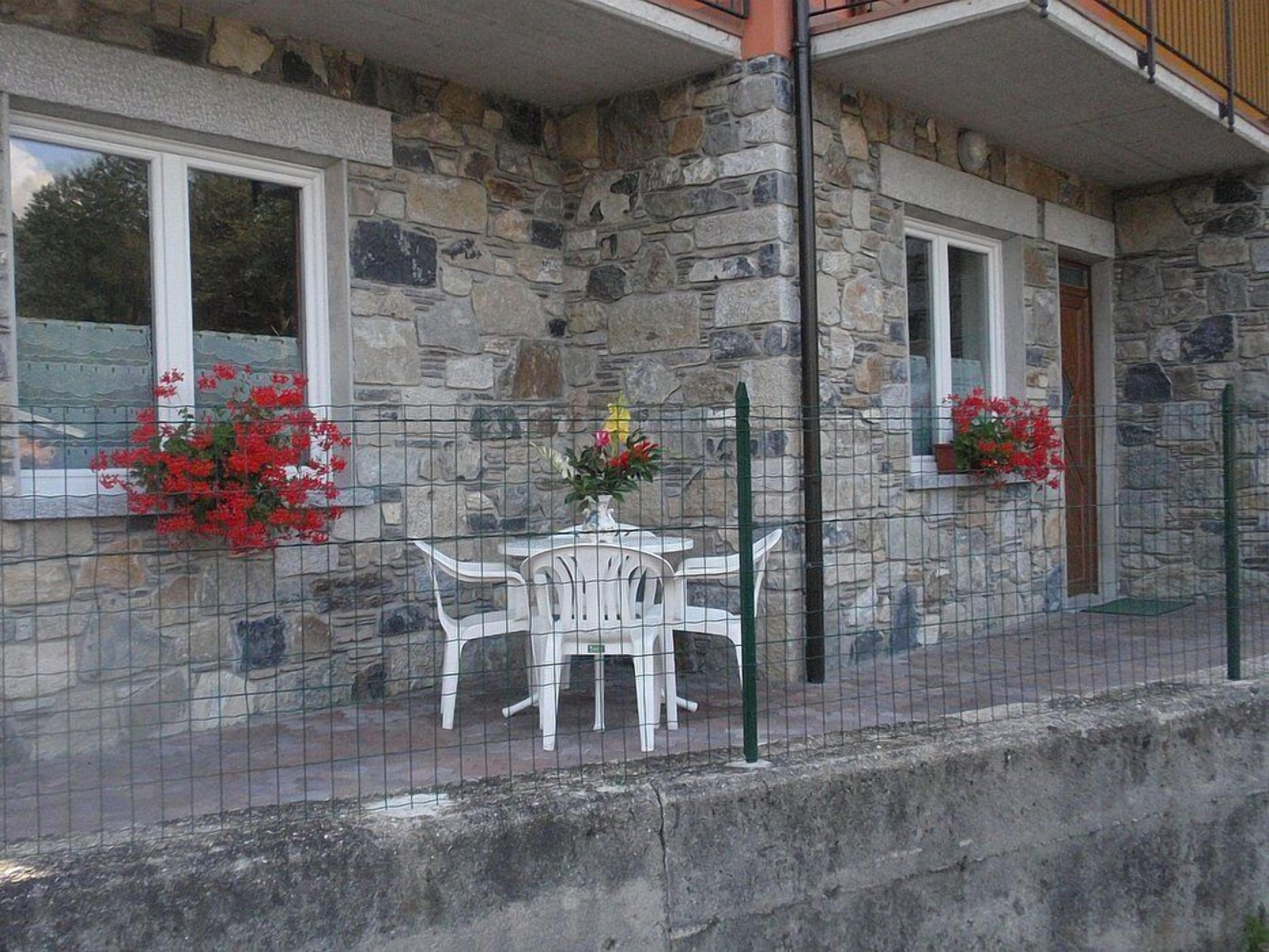 A stone building with a balcony, a table with chairs, and red flowers in window boxes.