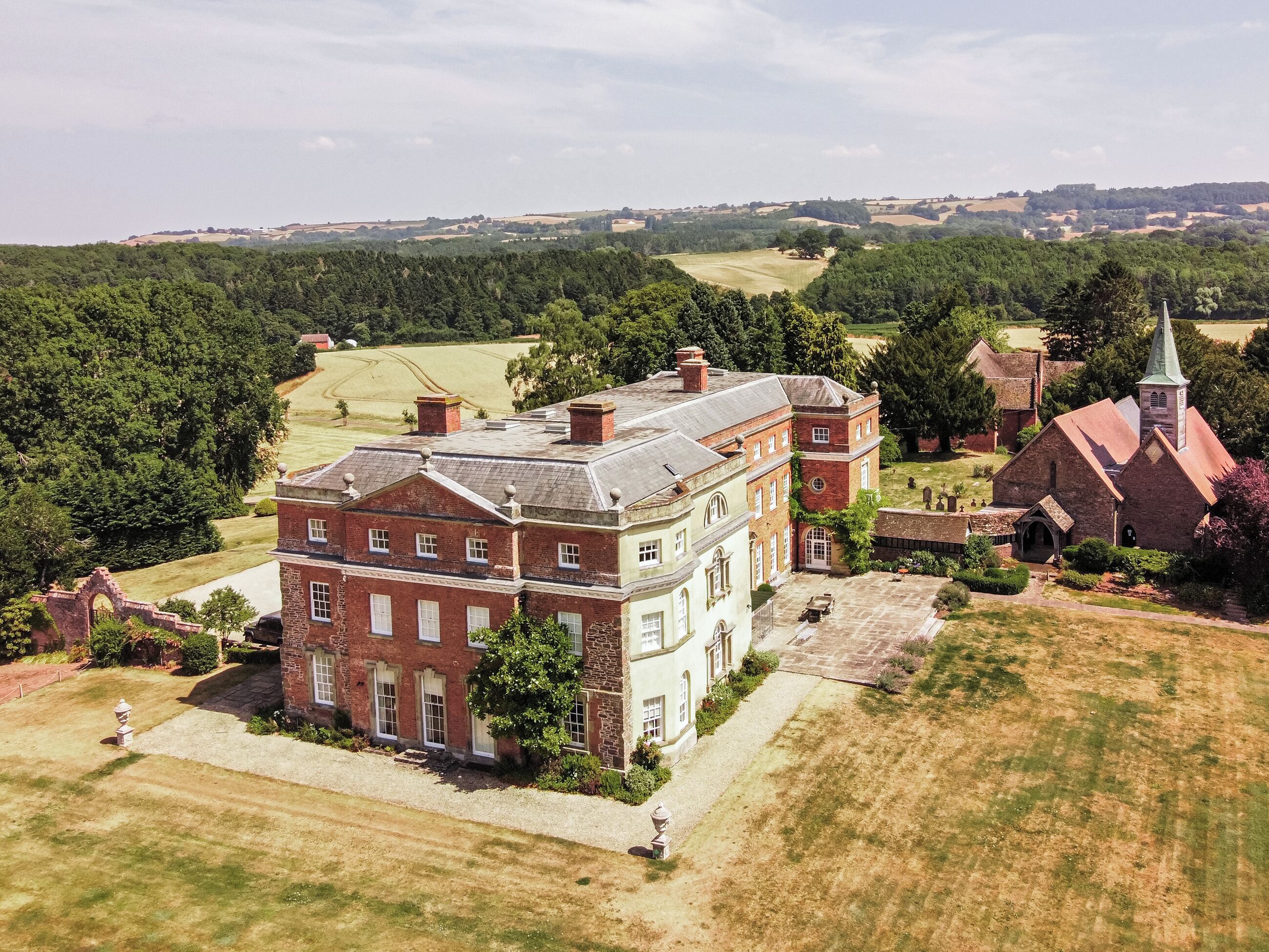 A large estate with a prominent red brick building, a church with a green spire, and expansive green fields.