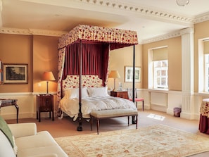Ein Bett mit Baldachin, ein Blumentapete-Kopfstück, ein Nachttisch mit Lampe, ein Sessel und ein großes Fenster, das natürliches Licht hereinlässt.