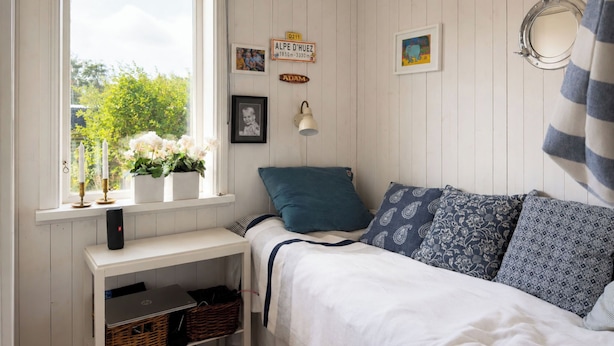 A bedroom with a bed, a window with white curtains, a bedside table with a lamp, and a wall-mounted mirror.
